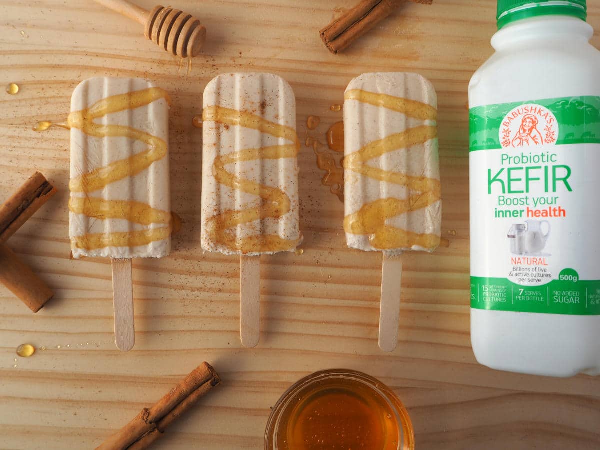 Single cinnamon, honey and kefir popsicle with honey drizzle and sprinkle of cinnamon, cinnamon stick, honey stick, honey bowl and milk kefir bottle on chopping board