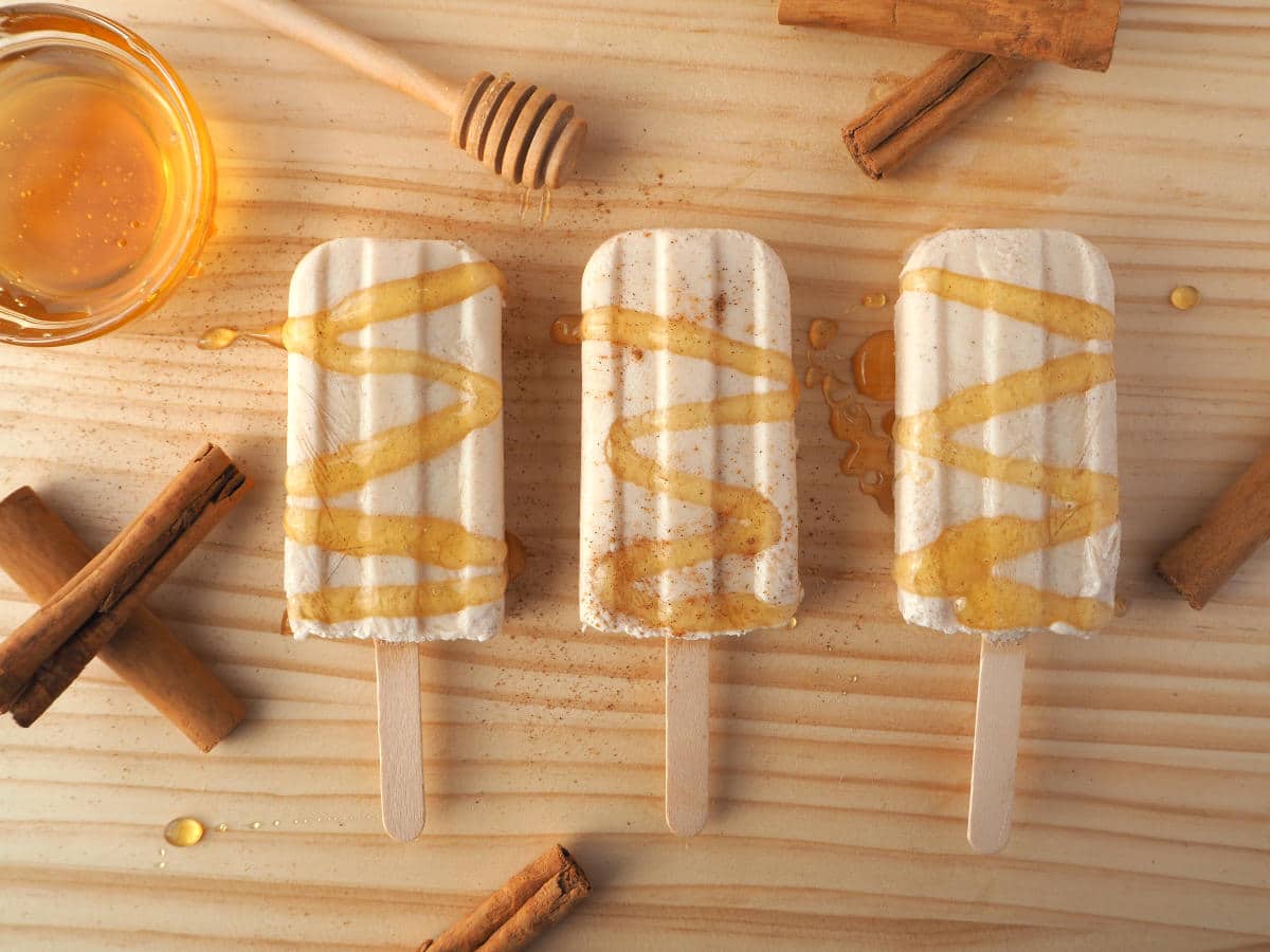 Row of three cinnamon, honey and kefir popsicles with honey drizzle and sprinkle of cinnamon, cinnamon stick, honey stick and honey bowl, on chopping board