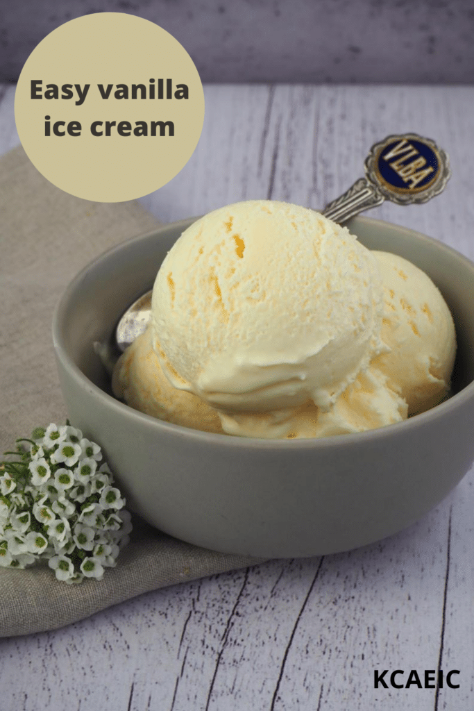 Scoops of vanilla ice ice cream in a grey bowl, with a vintage teaspoon, on a beige teatowl and a white wooded flood backgroud, with some small white flowers on the side, with text overlay, Easy vanilla ice cream and KCAEIC.