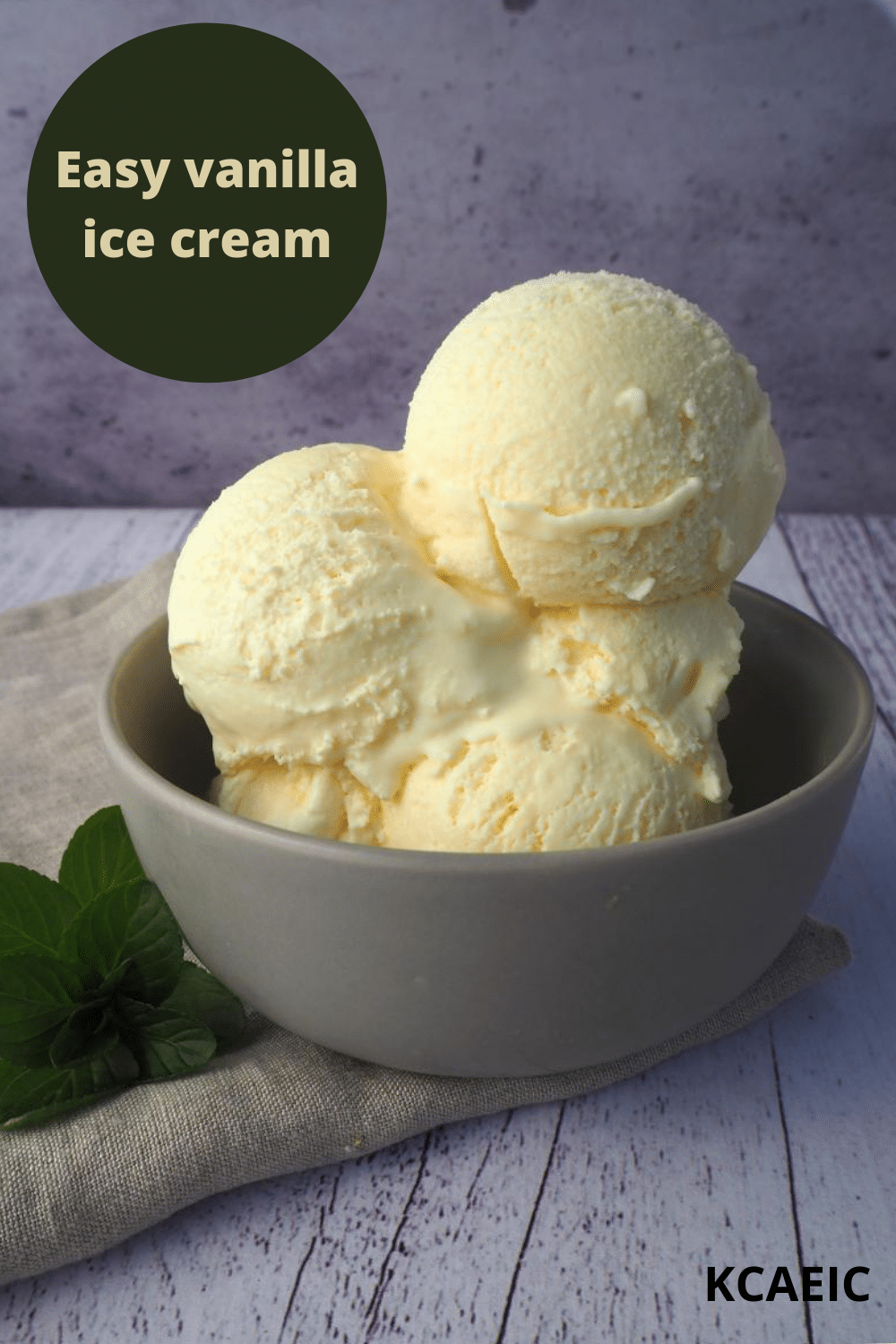 Scoops of vanilla ice ice cream in a grey bowl, with a vintage teaspoon, on a beige teatowl and a white wooded flood backgroud, with a sprig of mint on the side, with text overlay, Easy vanilla ice cream and KCAEIC.
