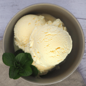 Top down view of a bowl of vanilla ice cream, in a grey bowl, with a spring of mint on the side in the bowl, on a beige tea towel and white wooded floor background.