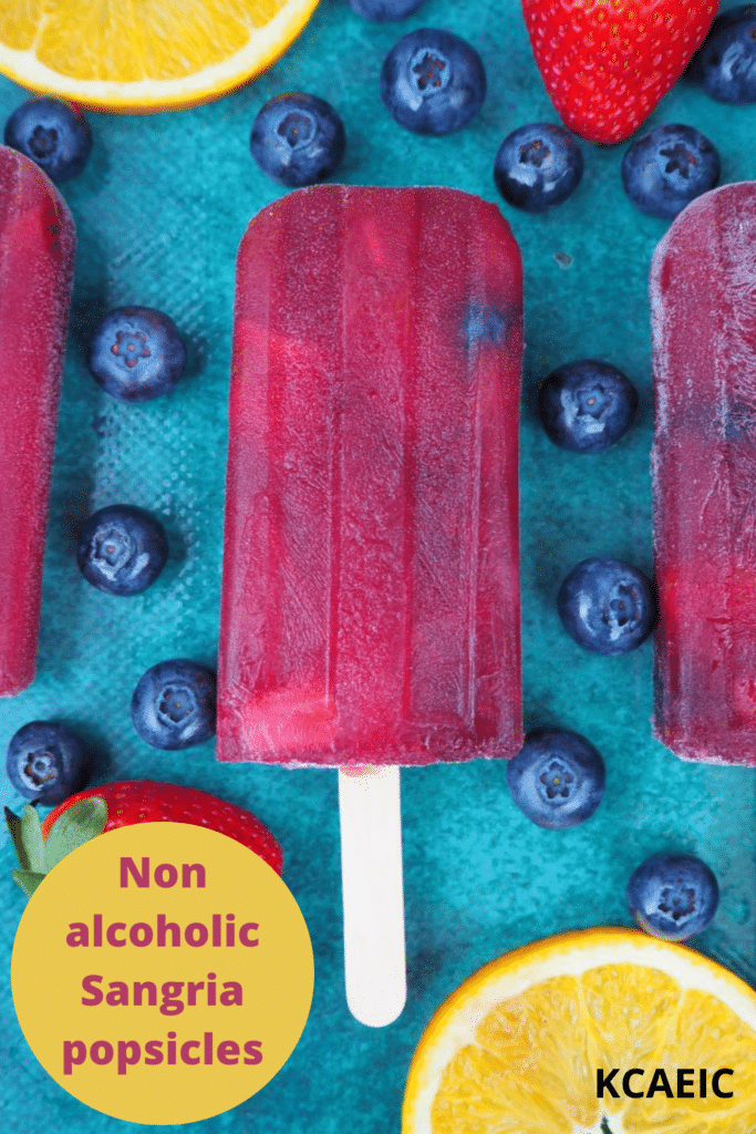 Close up of row of three non alcoholic sangria popsicle, on a green background, surrounded by strawberries, blueberries and orange slices, with text overlay, Non alcoholic sangria popsicles, KCAEIC