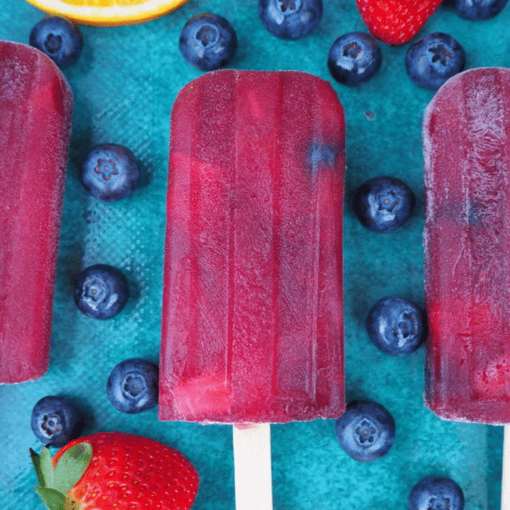 Close up of row of three non alcoholic sangria popsicle, on a green background, surrounded by strawberries, blueberries and orange slices