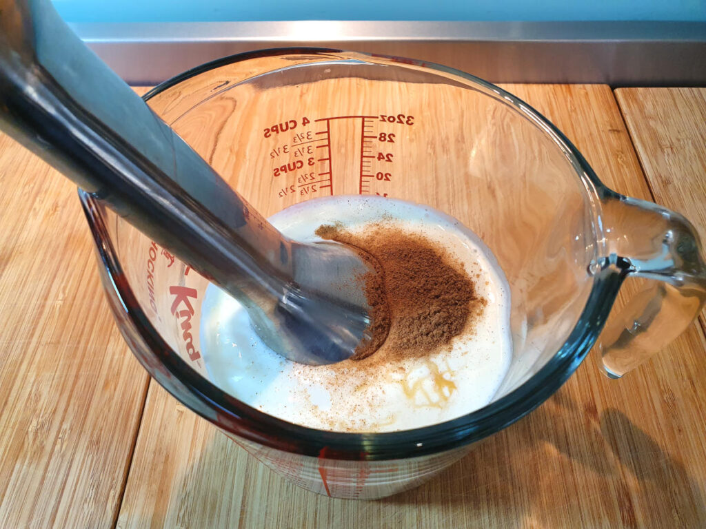 Blending cinnamon, honey and kefir mix in a glass jug with an immersion blender on a brown board