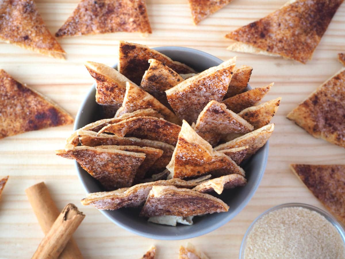 top down view of cinnamon sugar pita chips in bowl with pits chips around the sides, stacked cinnamon quills and a small glass bowl of sugar.