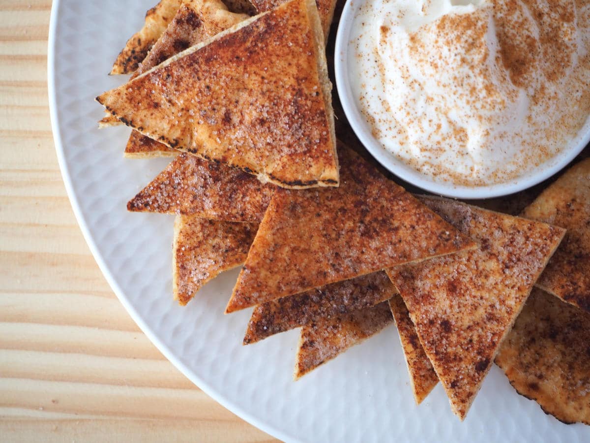 top down view of pile of cinnamon sugar pita chips on a white plate, with a bowl of cinnamon honey yogurt dusted with cinnamon sugar in the middle, on a wooded background.