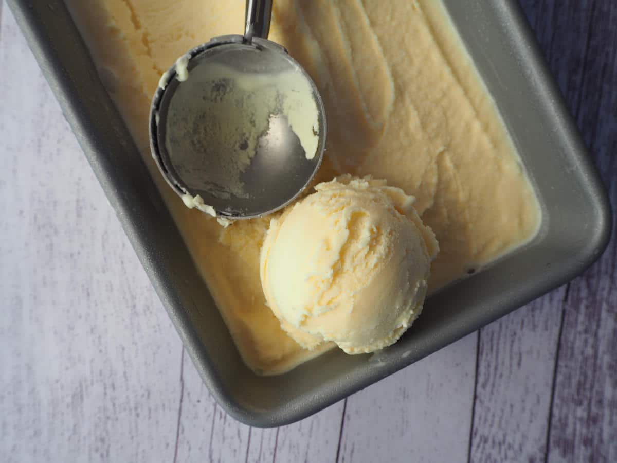 Top down view of a scoop of vanilla ice cream on top of ice cream, in a loaf pan, with an ice cream scooper, on a white wooded background.
