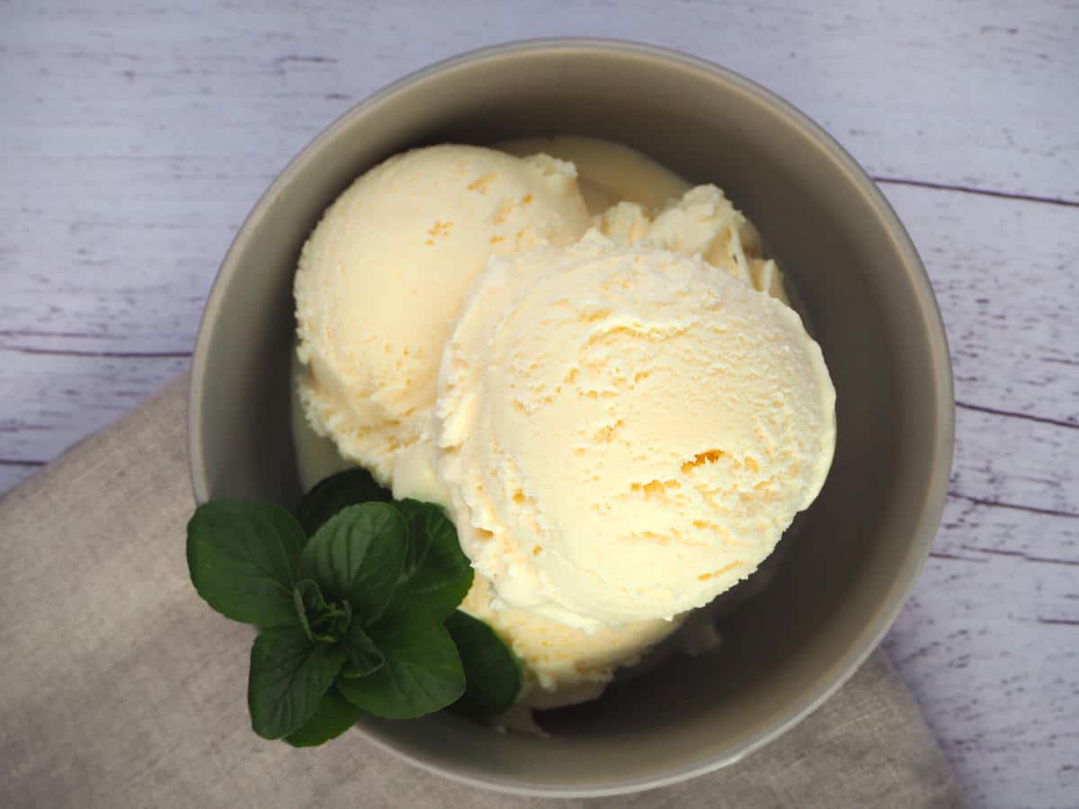 Top down view of a bowl of vanilla ice cream, in a grey bowl, with a spring of mint on the side in the bowl, on a beige tea towel and white wooded floor background.