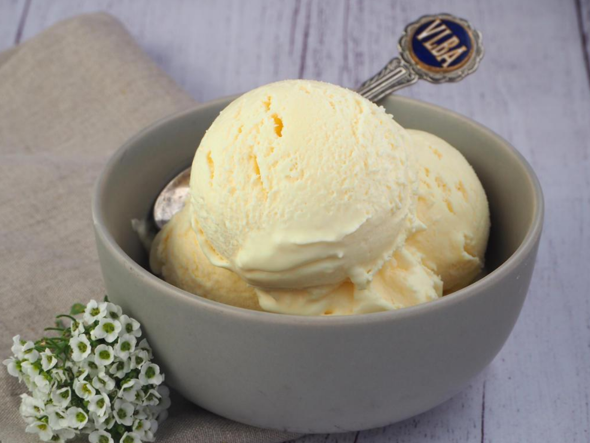 Scoops of vanilla ice ice cream in a grey bowl, with a vintage teaspoon, on a beige teatowl and a white wooded flood backgroud, with some small white flowers on the side.