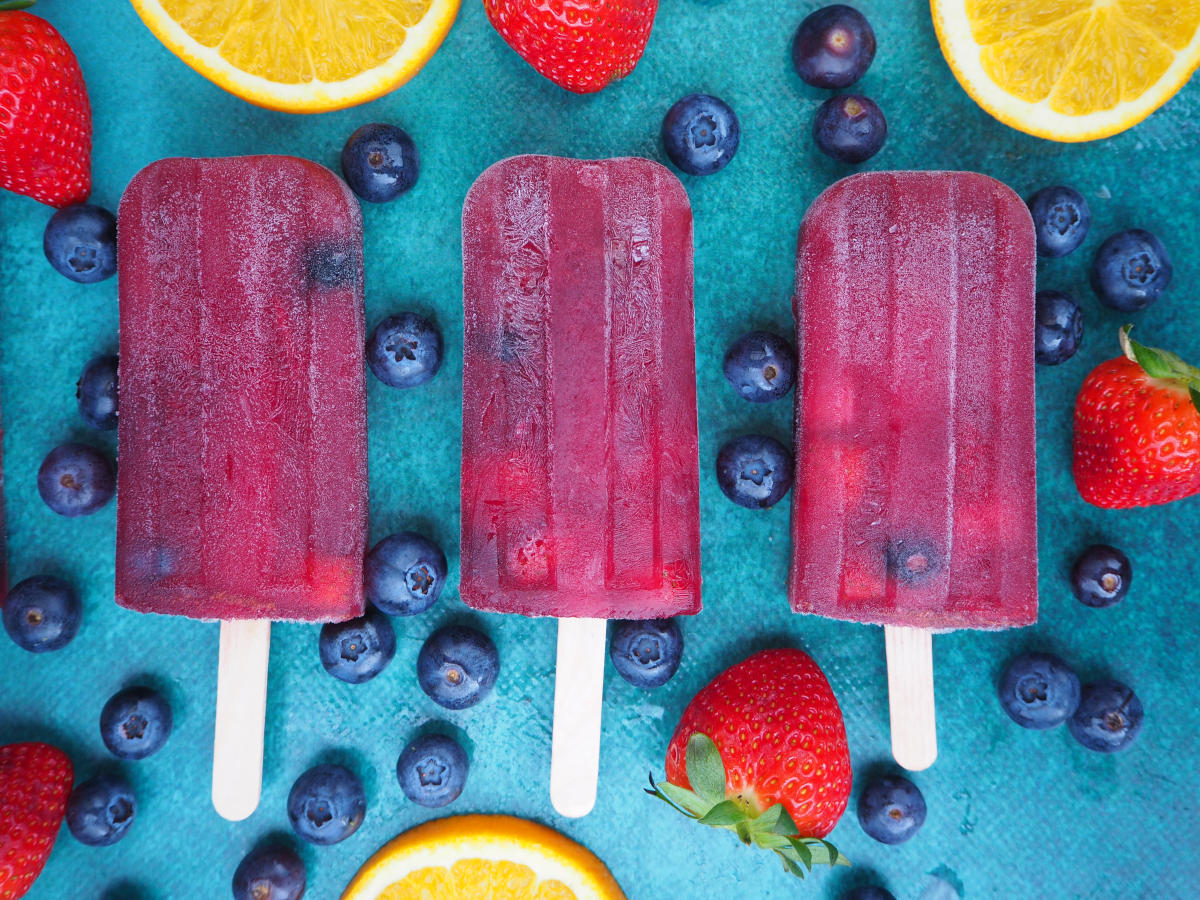 Close up of row of three non alcoholic sangria popsicle, on a green background, surrounded by strawberries, blueberries and orange slices