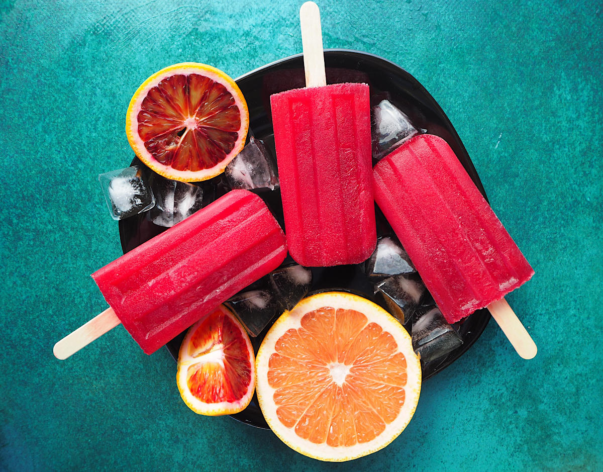 Ruby grapefruit and blood orange popsicle close up with orange and grapefruit slices and ice cubes on a black plate on a green background