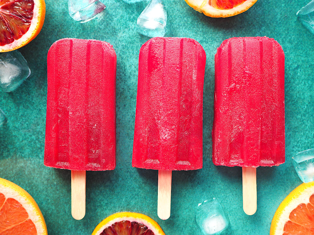 Row of three ruby grapefruit and blood orange popsicle close up with orange and grapefruit slices and ice cubes on green background