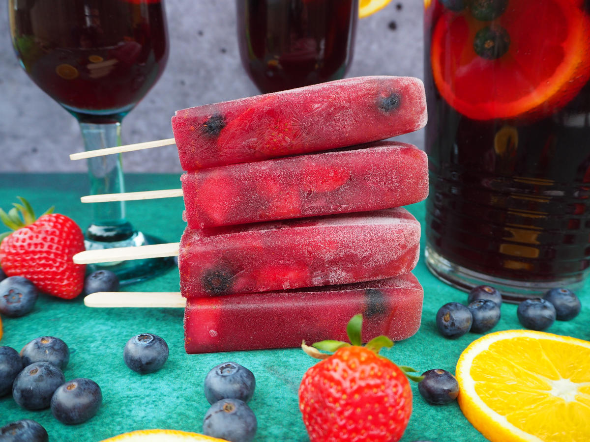 Close up of stack of four non alcoholic sangria popsicles side one, in front of a jug of sangria and two glasses of sangria, on a green background, with strawberries, blueberries and orange slices in the forground