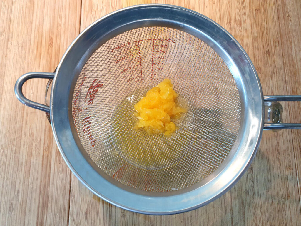 straining orange juice in a metal strainer over a jug on a chopping board