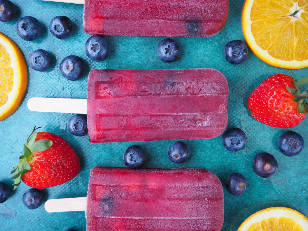 Close up of vertical row of three non alcoholic sangria popsicle, on a green background, surrounded by strawberries, blueberries and orange slices