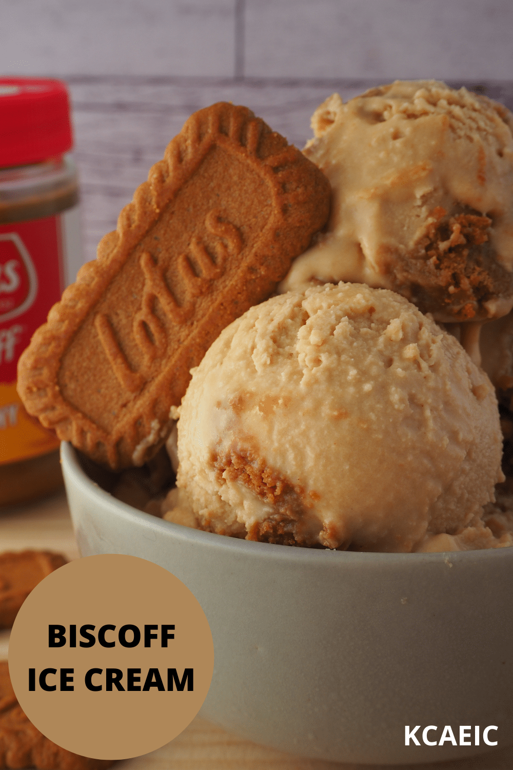 Side view of scoops of biscoff ice cream in grey bowl, with biscuit in ice cream on an an angle, with biscoff jar in the background, and text overlay, biscoff ice cream and KCAEIC.