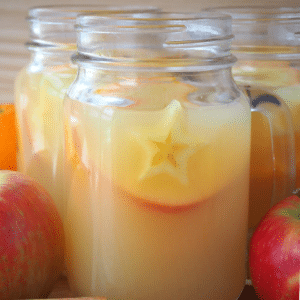 close up side view of a mason jar mug filled with mulled apple cider, with apples and spices on the side, on a brown board with a light brown background.