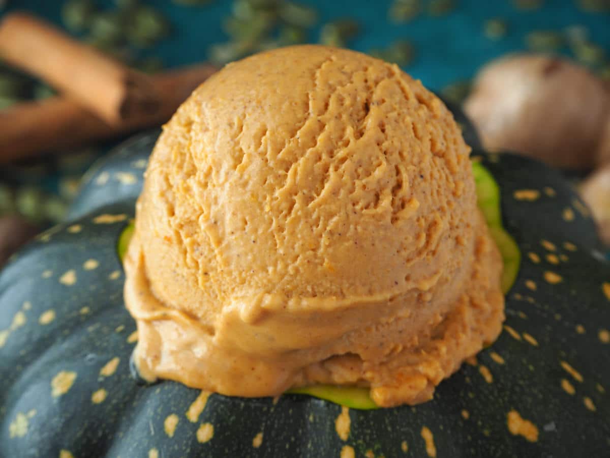 Close up side view of pumpkin spice ice cream scoop on a pumpkin, with blurry spices on a green background.