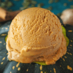 Close up side view of pumpkin spice ice cream scoop on a pumpkin, with blurry spices on a green background.