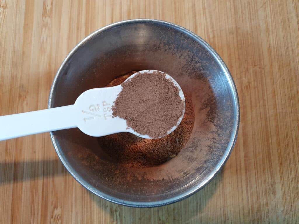 adding half a teaspoon of allspice to a small mental bowl, on a chopping board.