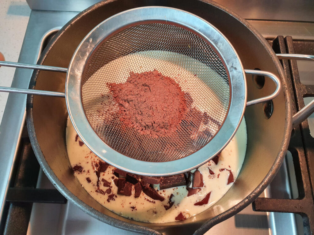 sifting cocoa powder over a pot on the stove with chocolate fudge sauce ingredients.