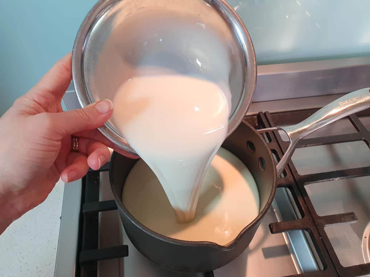 Pouring cream from a metal bowl into a small black pot on a gas stove.