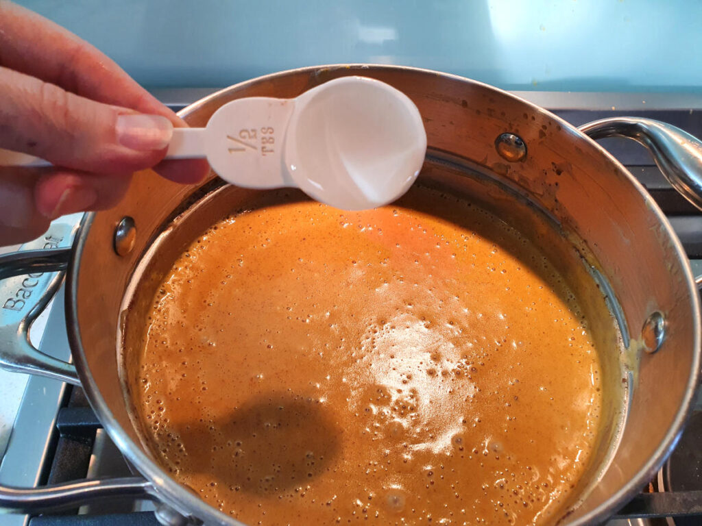 Adding half a tablespoon of glucose syrup to a double boiler with pumpkin spice ice cream mix on a gas stove.