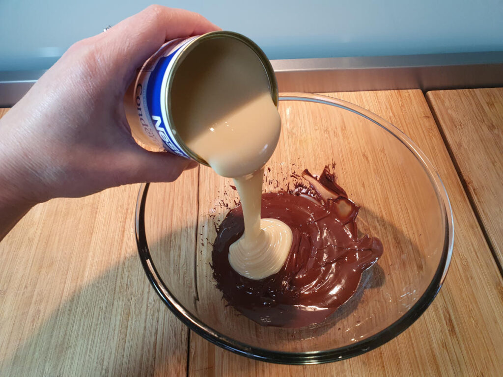 pouring in tin of sweetened condenced milk to melted chocolate in a glass bowl.