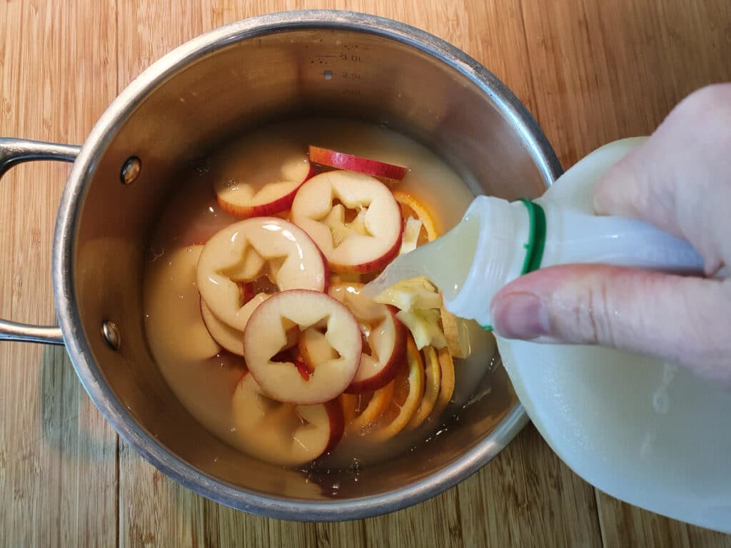 adding apple cider or cloudy apple juice to a pot containing sliced apples and oranges, on a chopping board.