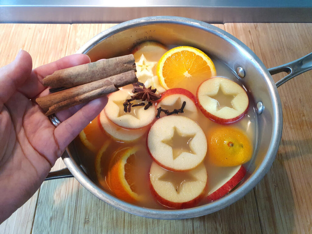 adding cinnamon sticks to pot containing sliced apples, oranges, cloves and star annise on a chopping board.