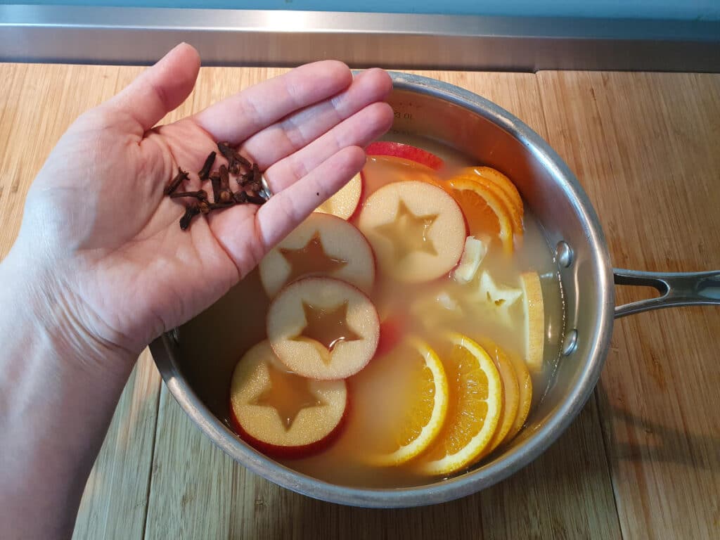 adding cloves to a pot containing sliced apples and oranges, and apple cider, on a chopping board.