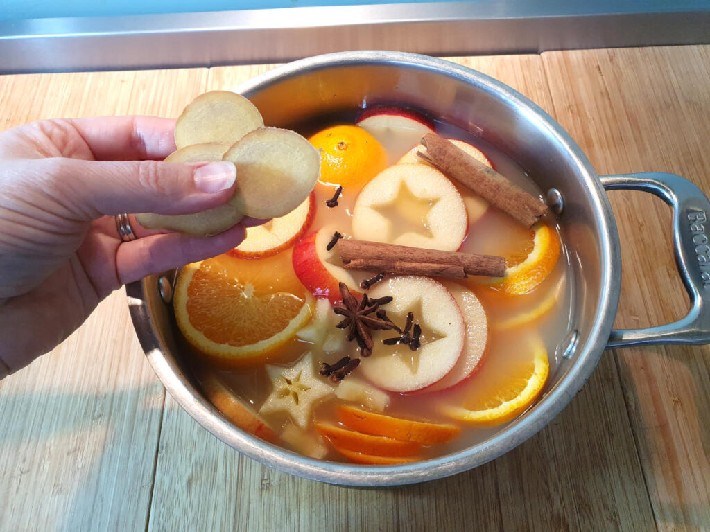 adding sliced ginger to a pot containing sliced apples and oranges, cloves, star annise and cinnamon sticks, on a chopping board.