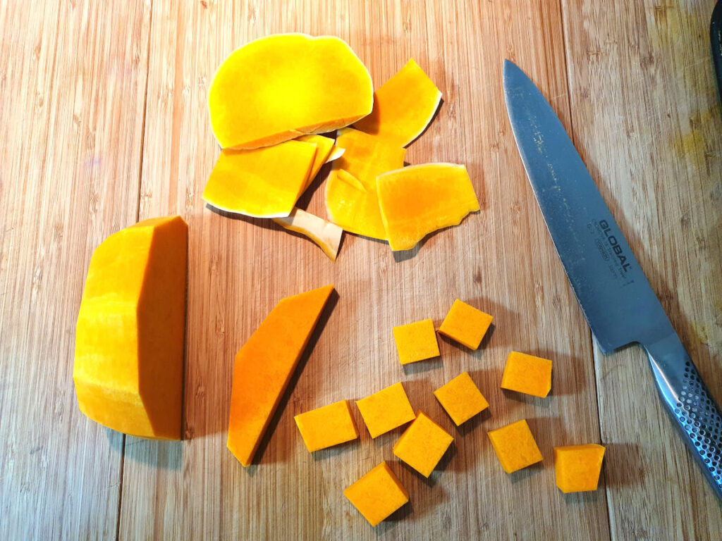 Top down showing pumpkin chopped up at various of chopping, with a large knife, on a chopping board.