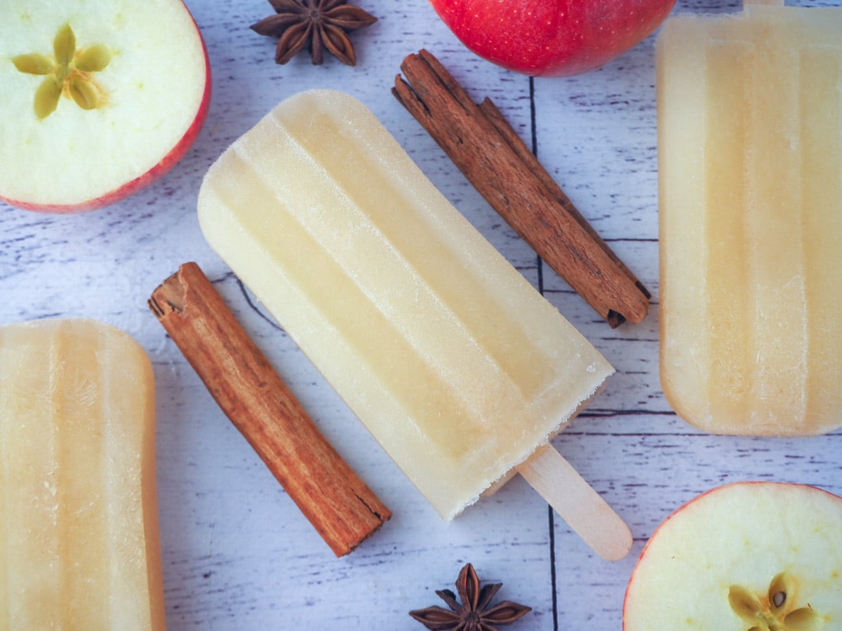 stack of three mulled apple cider popsicles in front of two glass mugs of mulled apple cider, with apple stars and star anise in the foreground, on a chopping board.