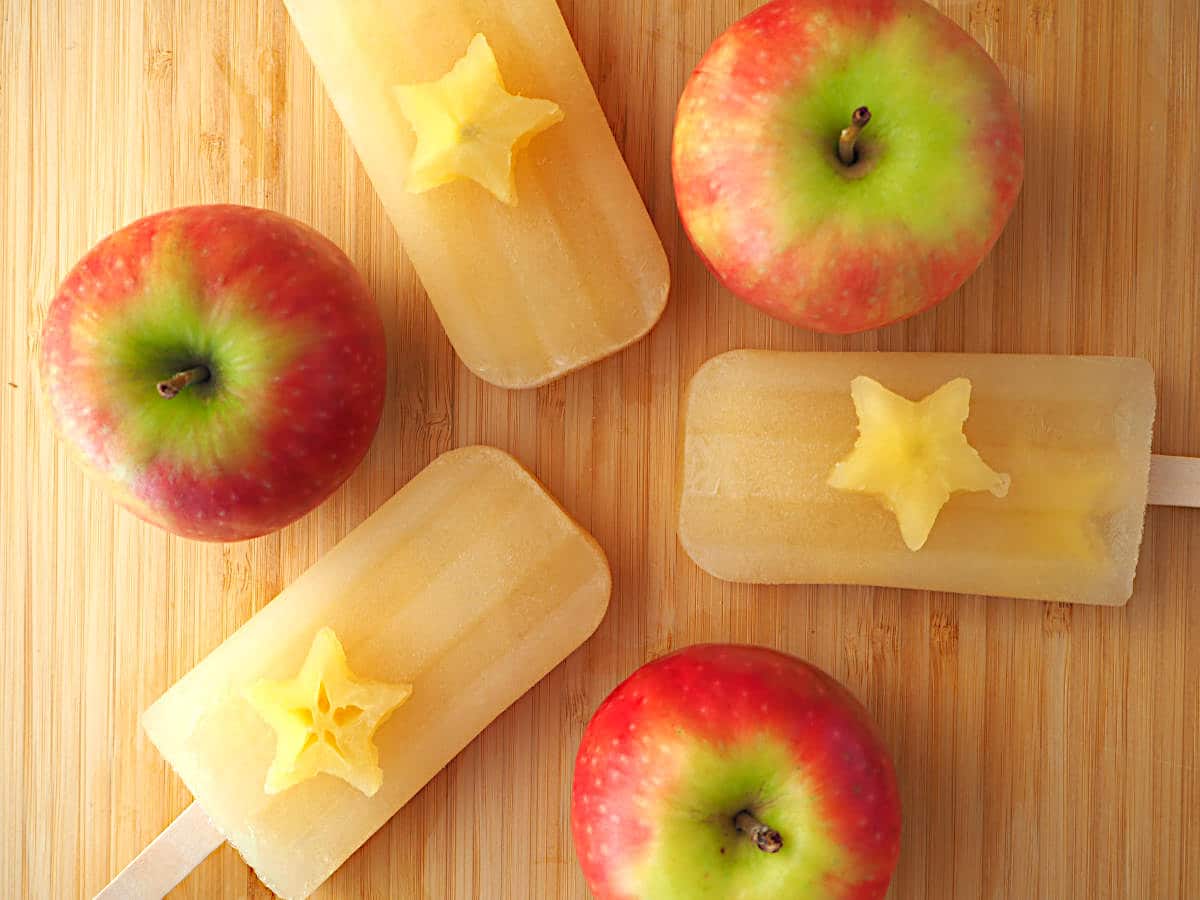 Three mulled apple cider popsicles in a trio all pointing in, with apple stars on top and apples in between, on a chopping board.