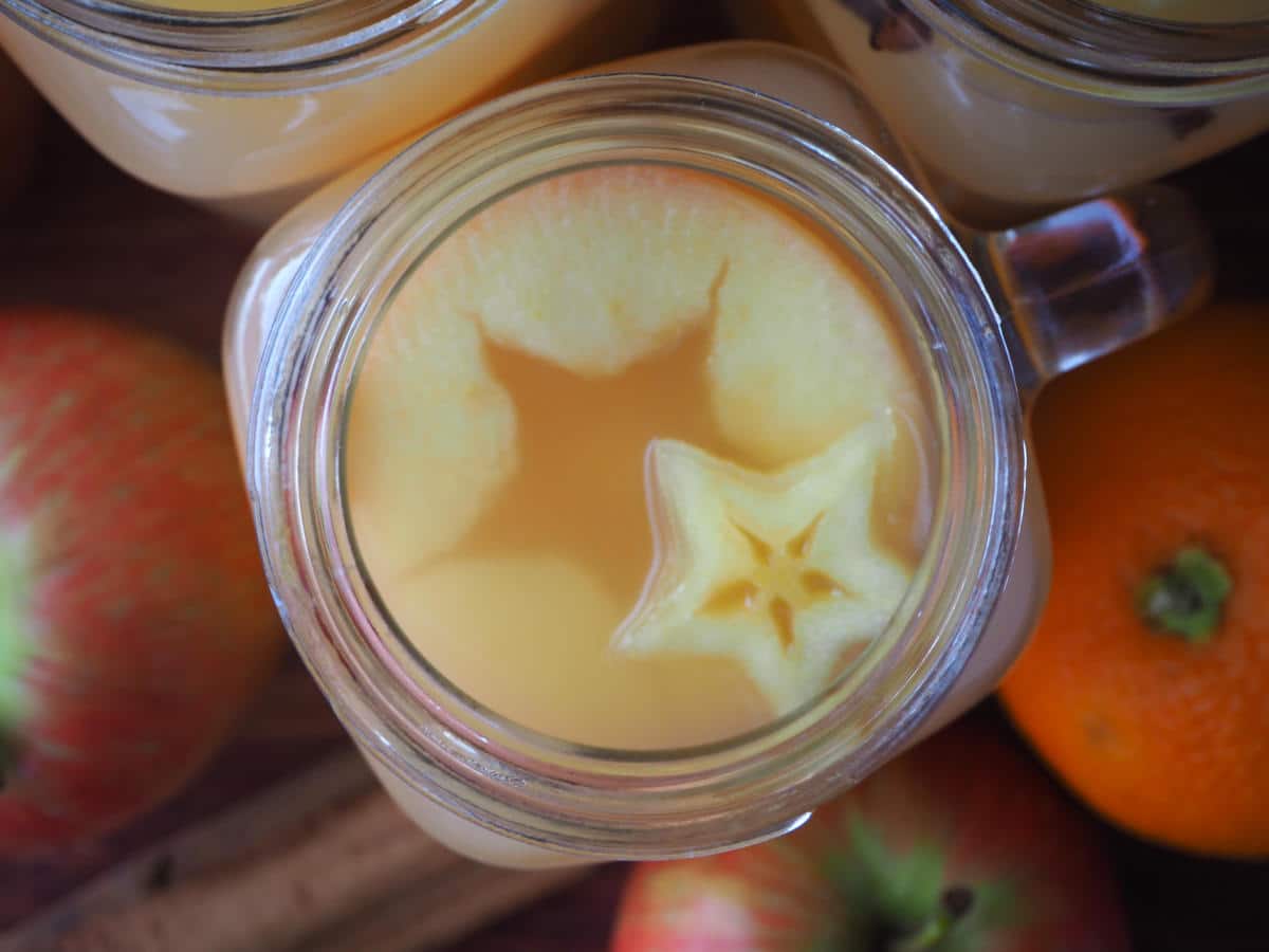 close up top down view of mulled apple cider showing star shapes, with apples, oranges and spices on the side.