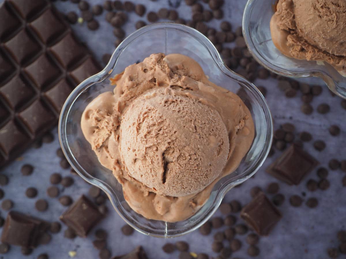 top down view of no churn chocolate ice cream in two sundae glasses, with chocolate pieces and chocolate chips, on a grey background.