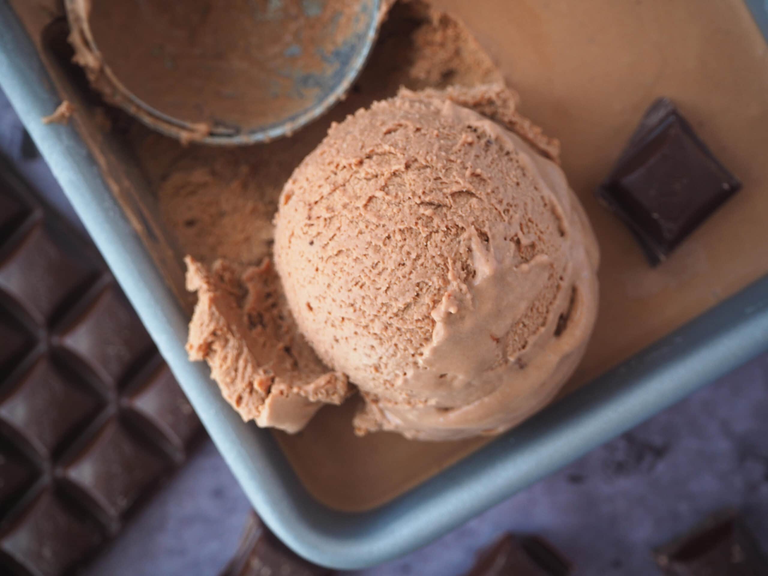 No churn chocolate ice cream 2 top down view of large scoop of no churn chocolate ice cream in pan, with ice cream scoop and pieces of chocolate on the side, on a grey background.