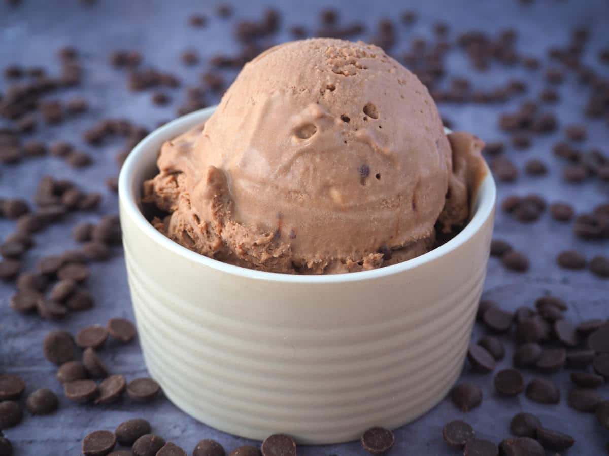 side view of large scoop of no churn chocolate ice cream in a small brown bowl, surrounded by chocolate chips, on a dark grey background.