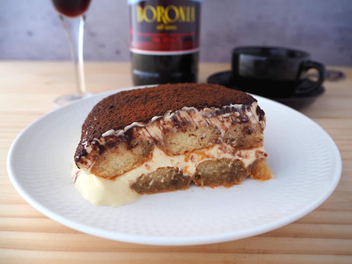 Side view of tiramisu on white plate with glass and bottle of marsala and black coffee cup with spoon.