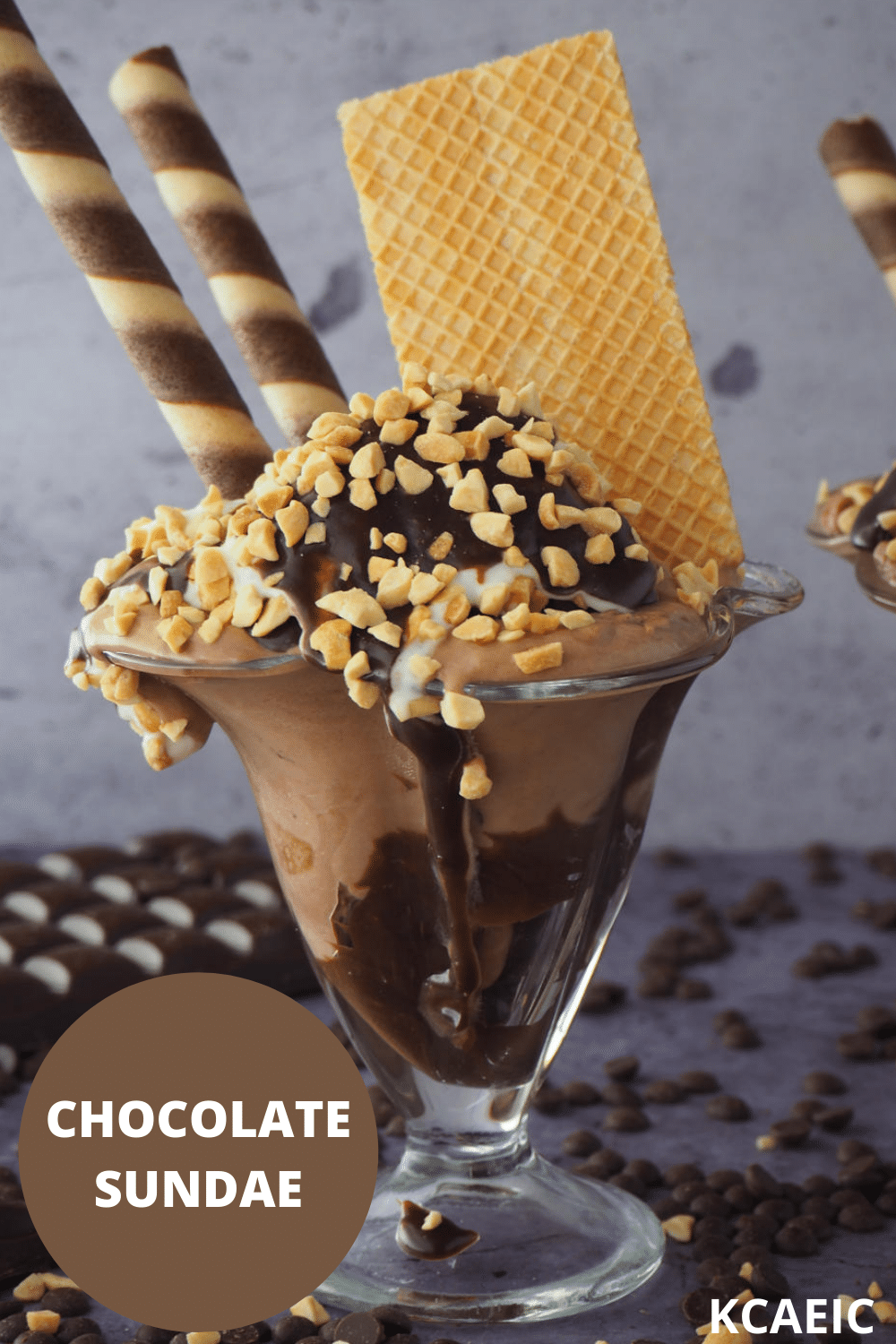 Side view of chocolate sundae in tall sundae glasses, with chocolate fudge sauce, chocolate ice cream, whipped cream, chopped nuts and chocolate and vanilla circular tube wafers, on a grey background, surrounded by chocolate pieces, chocolate chips and some chopped nuts, with text overlay, chocolate sundae and KCAEIC.