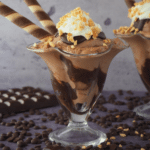 Side view of two chocolate sundaes in tall sundae glasses, with chocolate fudge sauce, chocolate ice cream, whipped cream, chopped nuts and chocolate and vanilla circular tube wafers, on a grey background, surrounded by chocolate pieces, chocolate chips and some chopped nuts.
