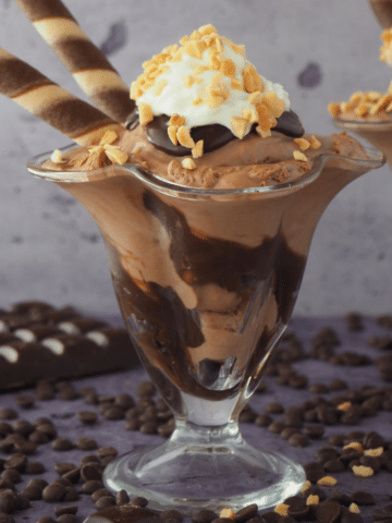 Side view of two chocolate sundaes in tall sundae glasses, with chocolate fudge sauce, chocolate ice cream, whipped cream, chopped nuts and chocolate and vanilla circular tube wafers, on a grey background, surrounded by chocolate pieces, chocolate chips and some chopped nuts.