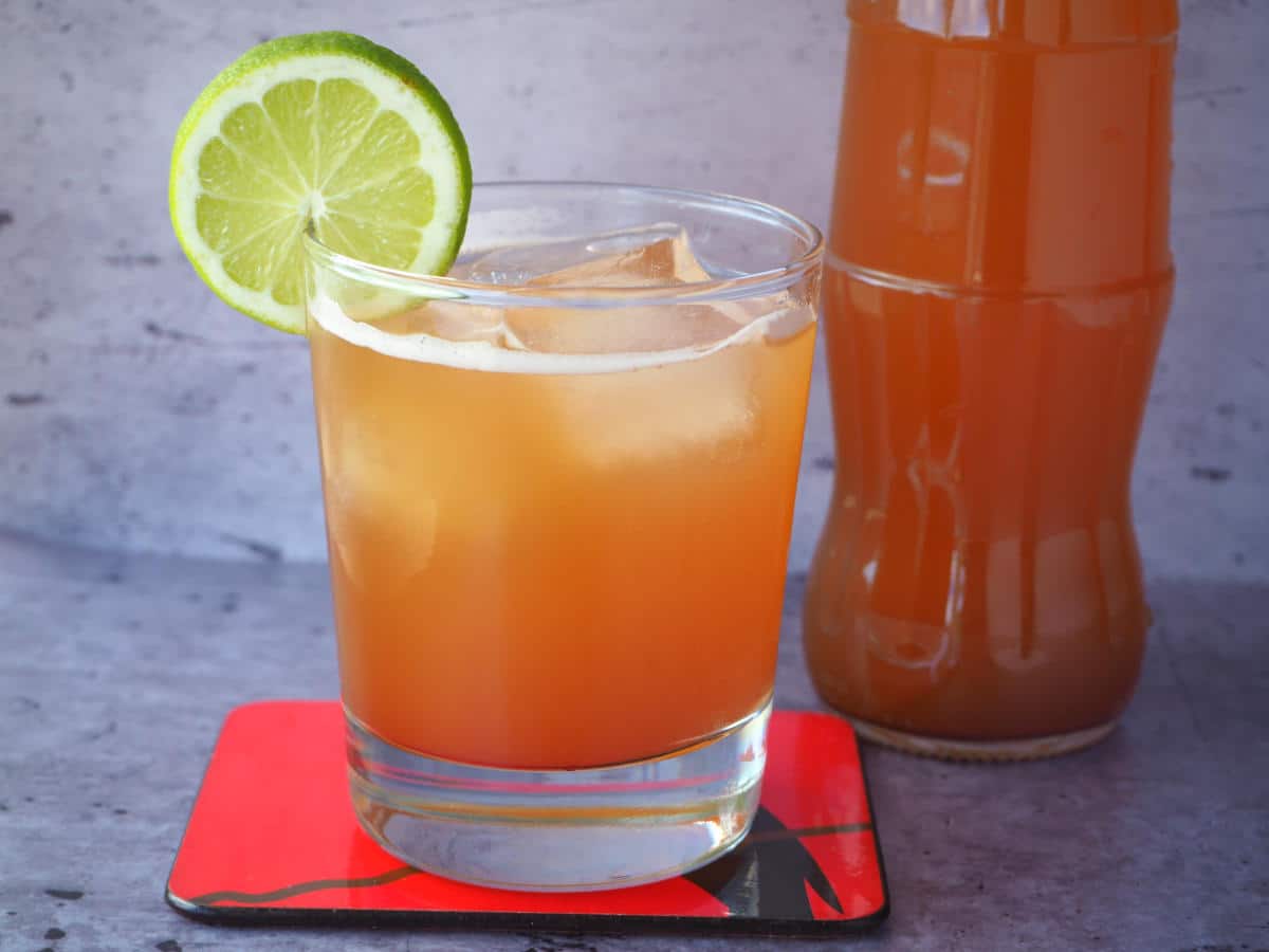 Glass of homemade cola with ice, lime circle garnish, red coaster under glass and bottle of cola beside it.