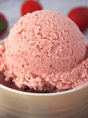 Close up side view of scoop of strawberry ice cream in a beige ramakin, with strawberries in the background.