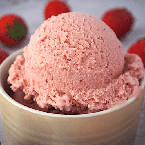 Close up side view of scoop of strawberry ice cream in a beige ramakin, with strawberries in the background.