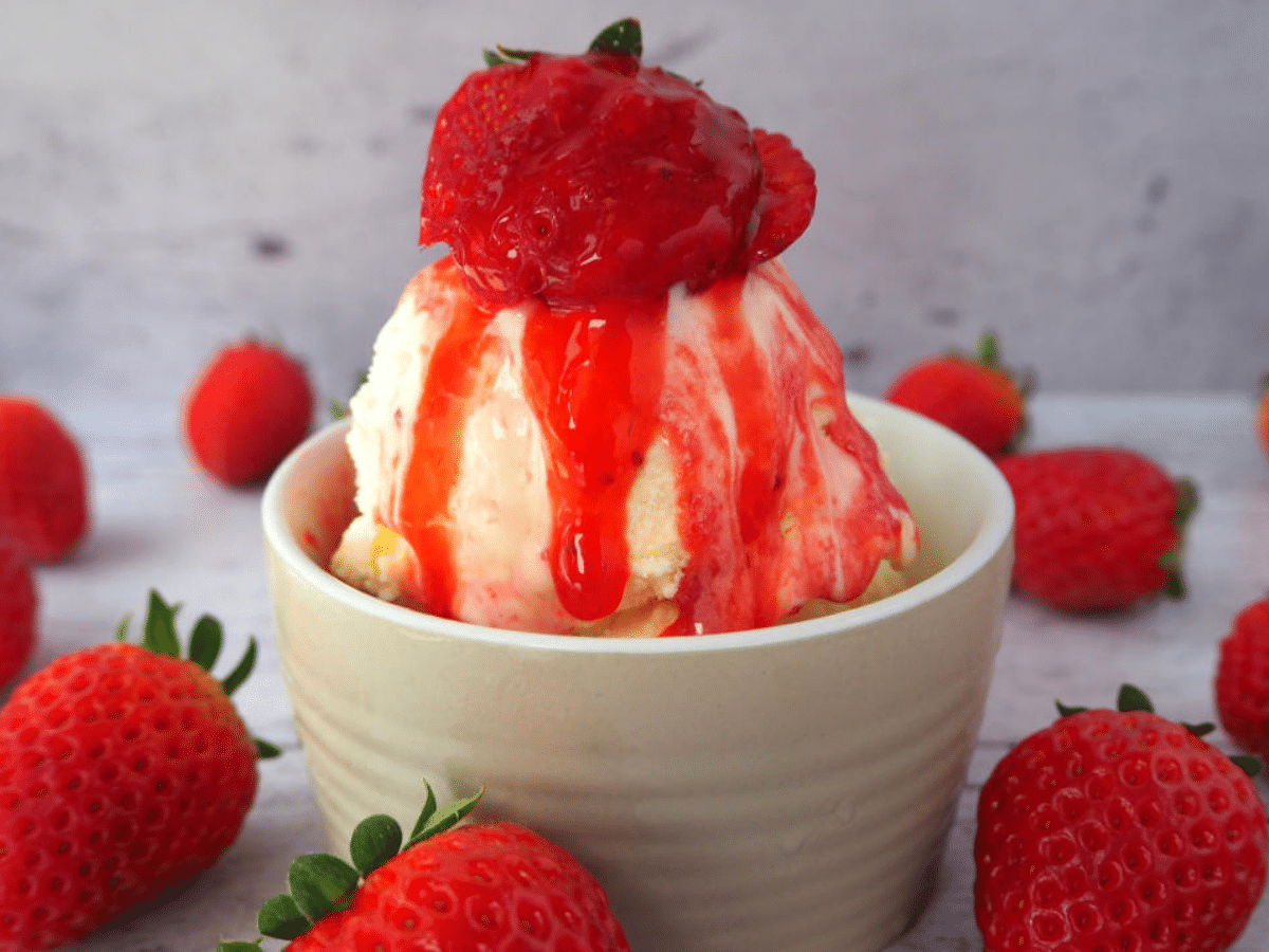Side view of vanilla ice cream with strawberry and strawberry sauce, with strawberries scattered around, on a white floorboard surface.