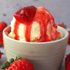 Close up side view of vanilla ice cream with strawberry and strawberry sauce, with strawberries scattered around, on a white floorboard surface