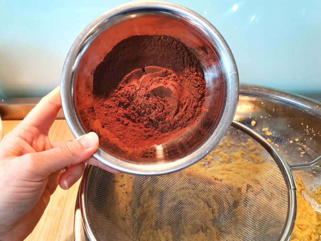 adding dutch cocoa powder to sieve sittnig on top of mixing bowl with beaten wet ingredients.