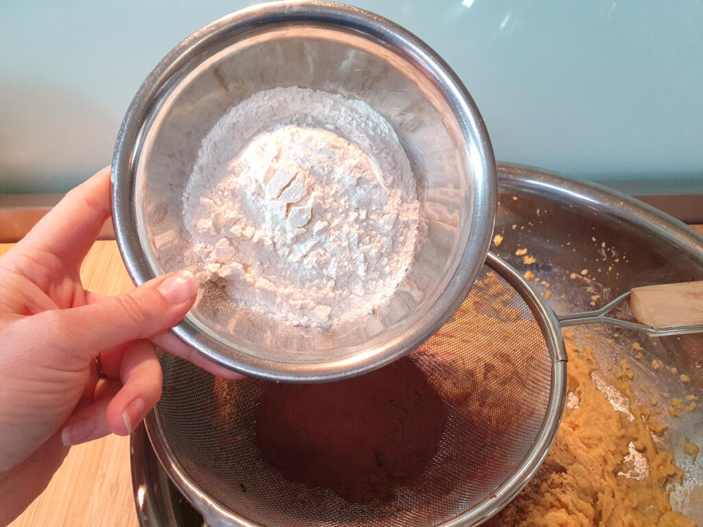adding flour to other dry ingredients in sieve sittnig on top of mixing bowl with beaten wet ingredients.